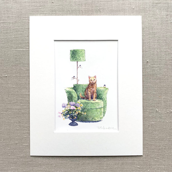 Matted artwork of a yellow cat on a green topiary chair with flowers and a lamp.