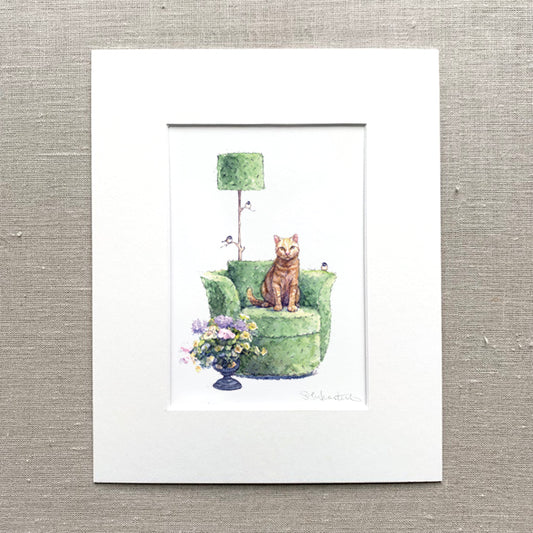 Matted artwork of a yellow cat on a green topiary chair with flowers and a lamp.
